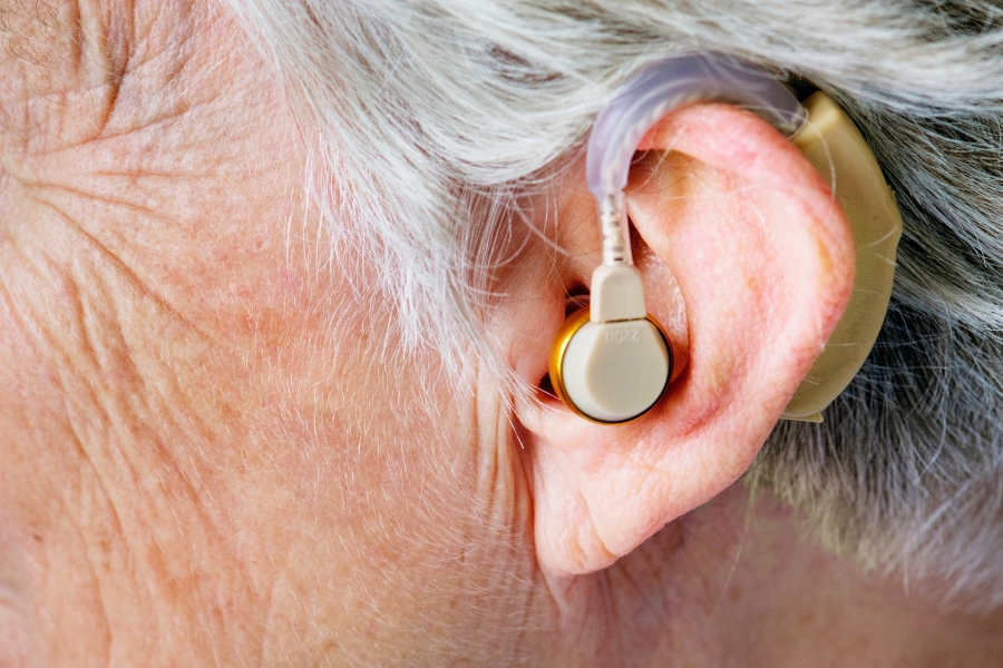 Close-up of an older person’s ear wearing a behind-the-ear hearing aid with a visible earpiece and cable, shown against grey hair and skin.