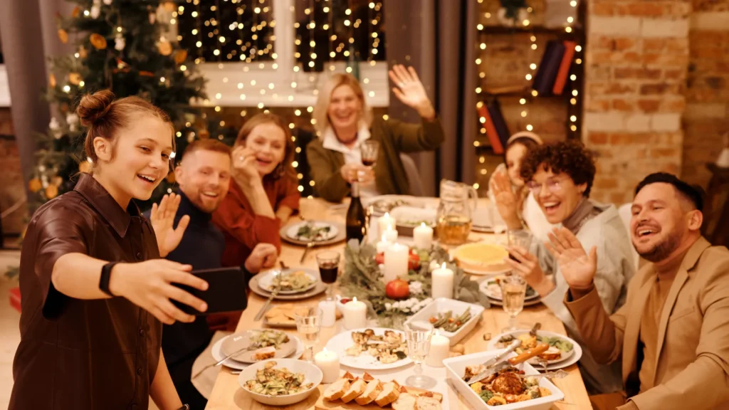 Family and friends gathered around a festive holiday dinner table, smiling and waving during a celebration.