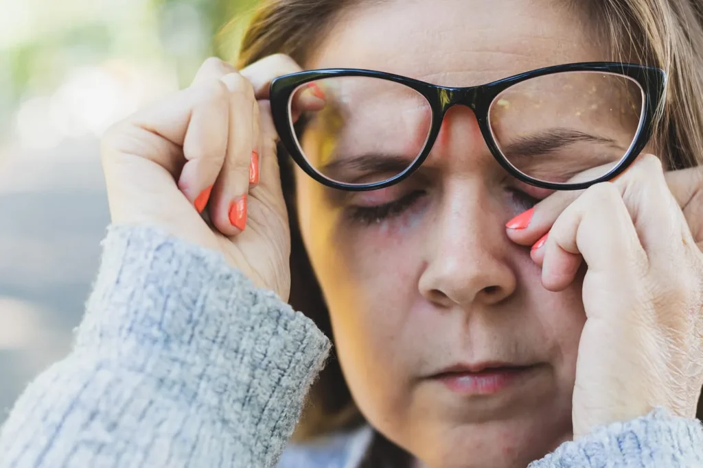Woman rubbing her eyes due to allergy symptoms such as itching and sinus irritation outdoors.